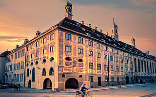 Abendlichtaufnahme des barocken Ursulinenklosters Landsberg am Lech mit bemalten Fassaden, Türmchen und Radfahrer auf Kopfsteinpflaster.