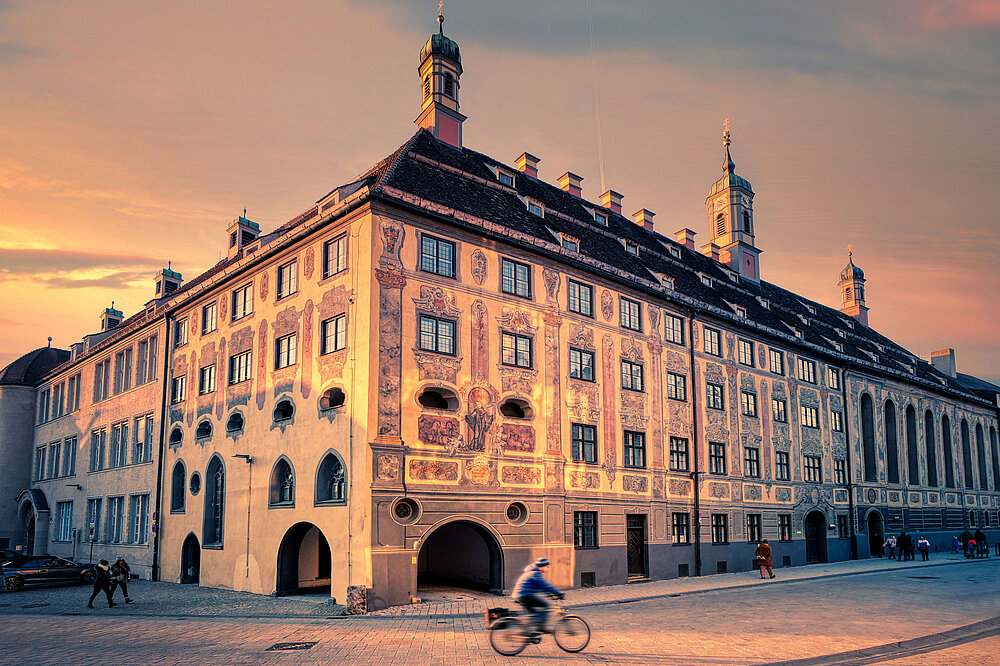 Abendlichtaufnahme des barocken Ursulinenklosters Landsberg am Lech mit bemalten Fassaden, Türmchen und Radfahrer auf Kopfsteinpflaster.