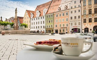 Gemütliche Café-Szene am Hauptplatz in Landsberg am lech: Apfeltarte und Kaffee auf dem Tisch, im Hintergrund historische Altstadthäuser, Mariensäule und bunte Fassaden unter blauem Himmel.