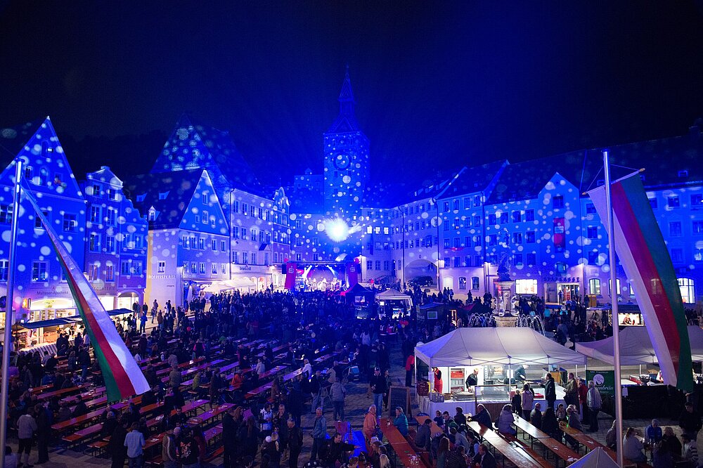 Nachtaufnahme des Lechstadtfests auf dem Hauptplatz Landsberg am Lech: Blaue Lichter und Projektionen auf Fachwerkhäusern und Kirchturm, belebter Markt mit Ständen, Bänken, Flaggen und Menschenmengen.