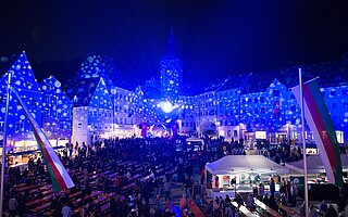 Nachtaufnahme des Lechstadtfests auf dem Hauptplatz Landsberg am Lech: Blaue Lichter und Projektionen auf Fachwerkhäusern und Kirchturm, belebter Markt mit Ständen, Bänken, Flaggen und Menschenmengen.