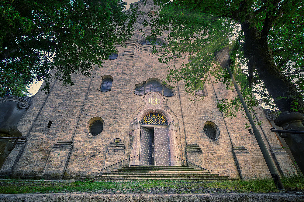 Frontalaufnahme der Heilig‑Kreuz‑Kirche Landsberg mit gotischem Portal, Rundfenstern und Bäumen vor dem Eingang.