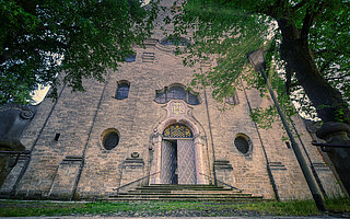 Frontalaufnahme der Heilig‑Kreuz‑Kirche (Malteserkirche) Landsberg am Lech mit gotischem Portal, Rundfenstern und flankierenden Bäumen.