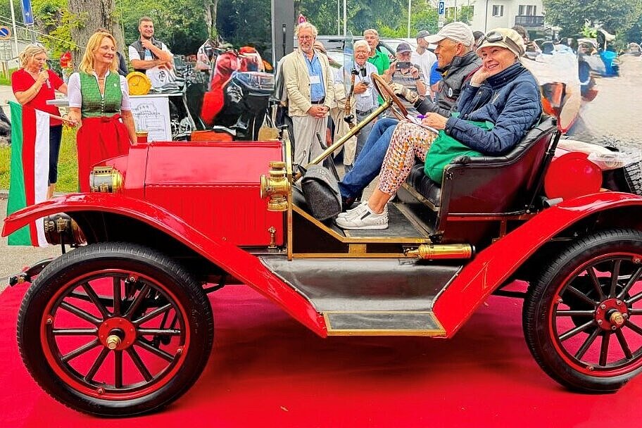 Bei der Herkomer-Konkurrenz: Paar sitzt lachend in rotem Oldtimer auf rotem Teppich in Landsberg am Lech, umgeben von jubelnder Menge.