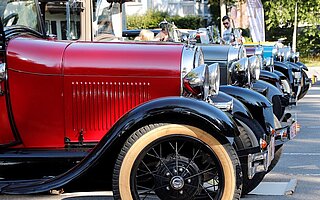 Reihe klassischer Oldtimer (rot, schwarz, beige) vor Landsberger Häusern: Vorkriegsautos mit gelben Speichenrädern und Zuschauern am Straßenrand.