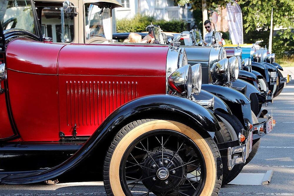 Reihe klassischer Oldtimer (rot, schwarz, beige) vor Landsberger Häusern: Vorkriegsautos mit gelben Speichenrädern und Zuschauern am Straßenrand.
