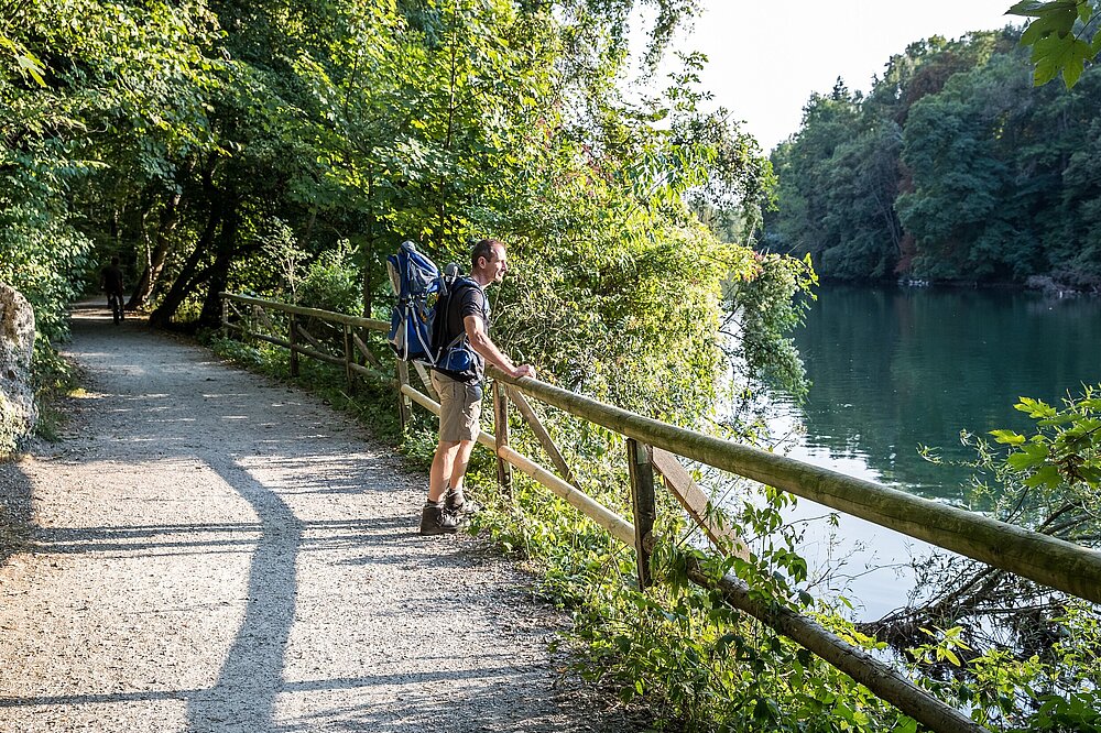 Wanderer mit Rucksack lehnt an Geländer auf Pfad durch Grünanlage mit Ausblick auf Lech-Fluss im Wildpark Pössinger Au bei Landsberg am Lech – Wandern Lechpark Bayern (CC BY-SA Klinger)