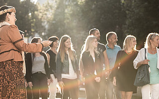 Eine Frau in traditioneller bayerischer Tracht führt begeisterte Besucher durch die historische Altstadt von Landsberg am Lech. Authentische Führung mit Insider-Geschichten und Leidenschaft für die Stadtgeschichte.