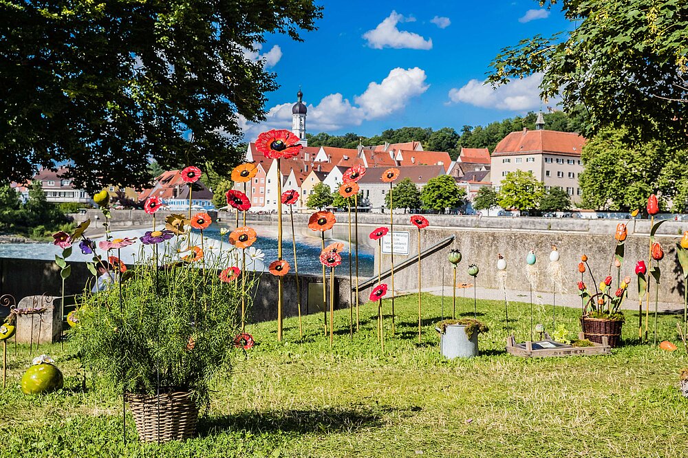 Bunte Mohnblumen-Skulpturen aus Metall in Orange, Rot und Violett vor der historischen Altstadt Landsberg am Lech mit Kirchturm und Lechbrücke – Kunstinstallatioxfer.