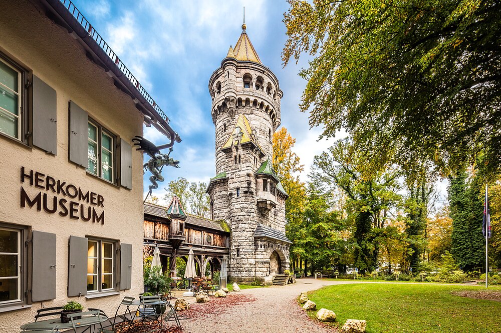 Herkomer-Museum am Mutterturm Landsberg: Schild 'HERKOMER MUSEUM', romantischer Steinturm mit Fachwerkgebäude, gelbe Bäume und Kiesweg