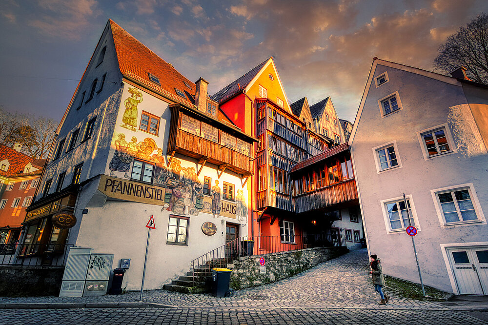 Das Foto zeigt das farbenfrohe Pfannenstiel-Haus (früher Obstgeschäft Pfannenstiel) im Hexenviertel von Landsberg am Lech bei Sonnenuntergang mit "Pfannenstiel" Schild und einer Person auf der Treppe.