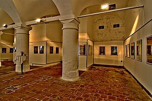 Historische Säulenhalle Landsberg: Beleuchtete Rundbögen und Säulen umgeben Ausstellungswände mit Gemälden, gedeckter Tisch und Steinfußboden – Kunstraum mit Backsteinarchitektur.