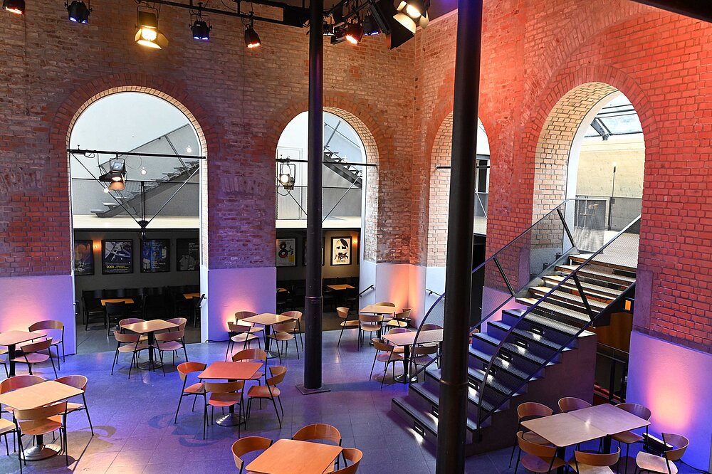 Modern gestaltetes Theaterfoyer in Backsteinbau Landsberg: Bunte Beleuchtung, Holztische, Treppen und hohe Rundbögen mit Blick auf die Stadt.