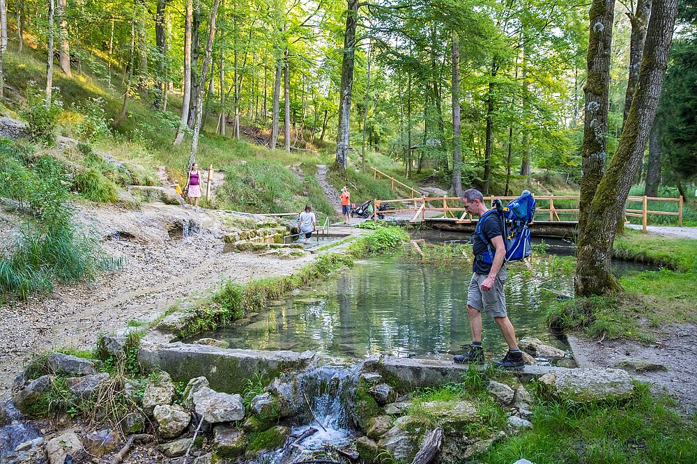 Wanderer mit Rucksack steht am Kneippbecken mit klarem Wasserbecken und Fußbad in der Natur des Wildparks Pössinger Au Landsberg am Lech – Wandern Lechpark Bayern mit Wellness (CC BY-SA Klinger)