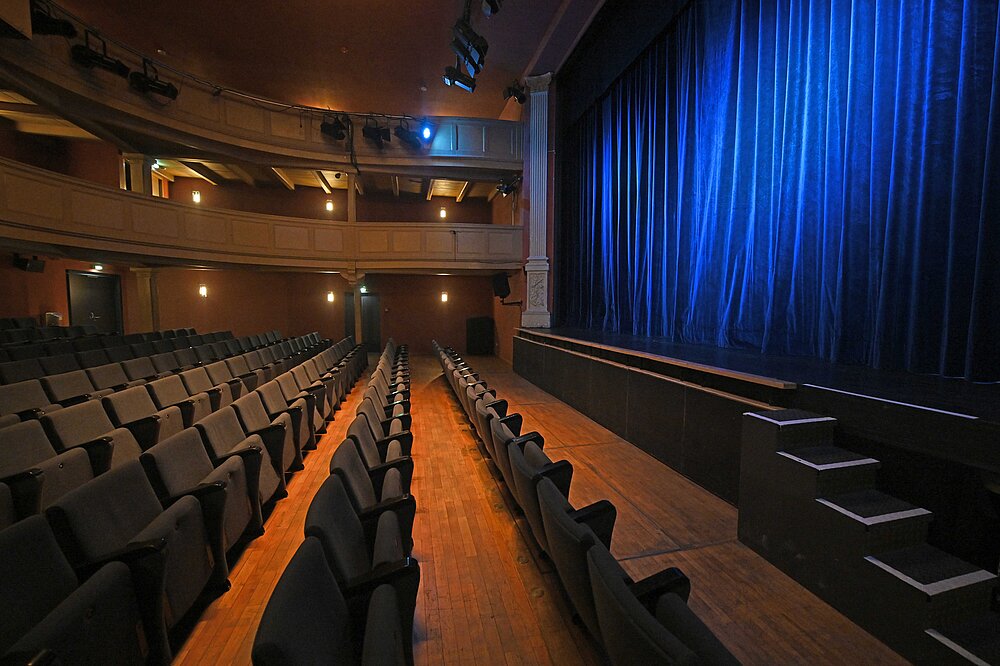 Stadttheater Landsberg: Klassischer Zuschauerraum mit Balkon, Holzboden und blauem Vorhang auf der Bühne – beleuchtet für Aufführungen.