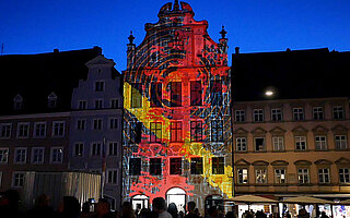Fassade des historischen Rathauses Landsberg am Lech bei der Langen Kunstnacht: Farbige Lichtprojektionen in Rot, Gelb und Blau leuchten in der Dunkelheit, davor Menschenmenge am Hauptplatz.