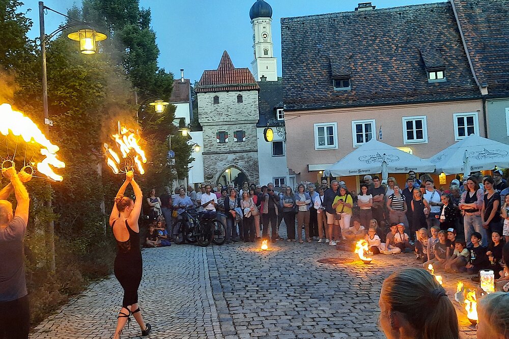 Feuerkünstler:innen performen bei der Langen Kunstnacht in Landsberg vor dem Bayertor, umringt von Zuschauern auf dem Kopfsteinpflaster, lodernde Flammen erhellen den Abendhimmel.