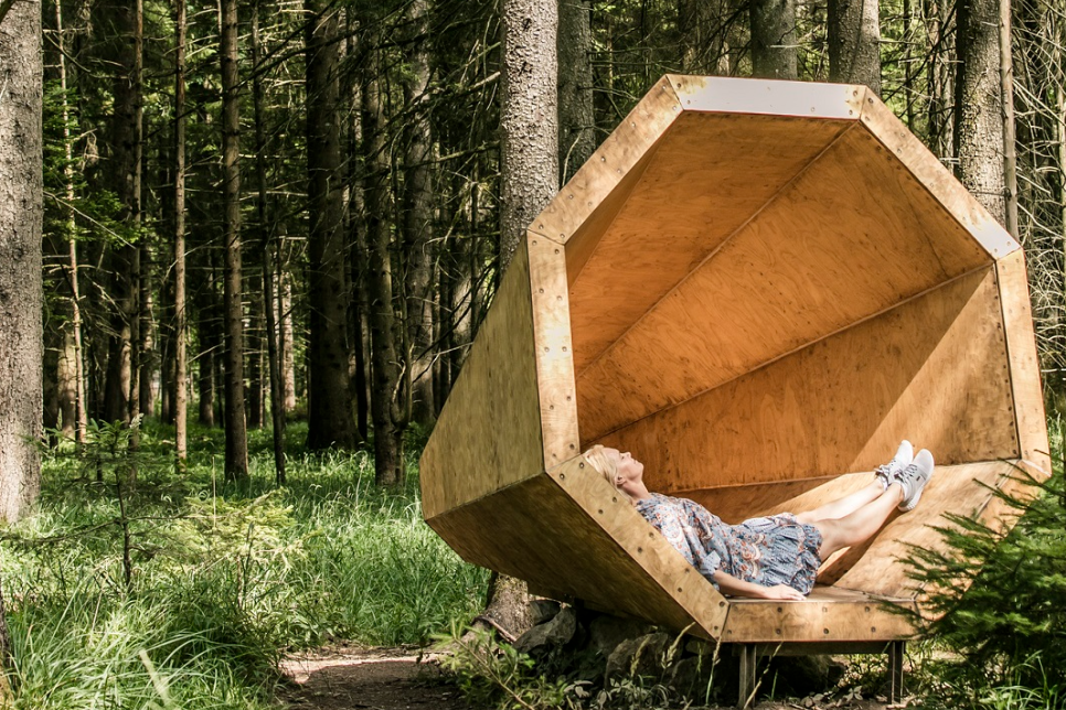 Frau liegt entspannt in einem achteckigen, offenen Holzpodest (Lauschplatz) im dichten Wald, umgeben von Fichten, Moos und jungen Bäumen – erholsame Naturarchitektur.