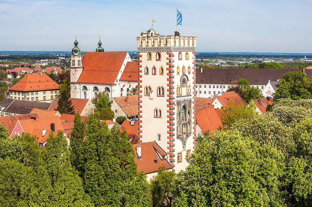 Luftaufnahme des Bayertors in Landsberg am Lech: Gotisches Stadttor aus 1425 mit weiß-rotem Turm, bayerischer Flagge, Altstadtdächern, Kirchen und Lechpanorama – Wahrzeichen Bayerns, 36m hoch, Aussichtsturm.