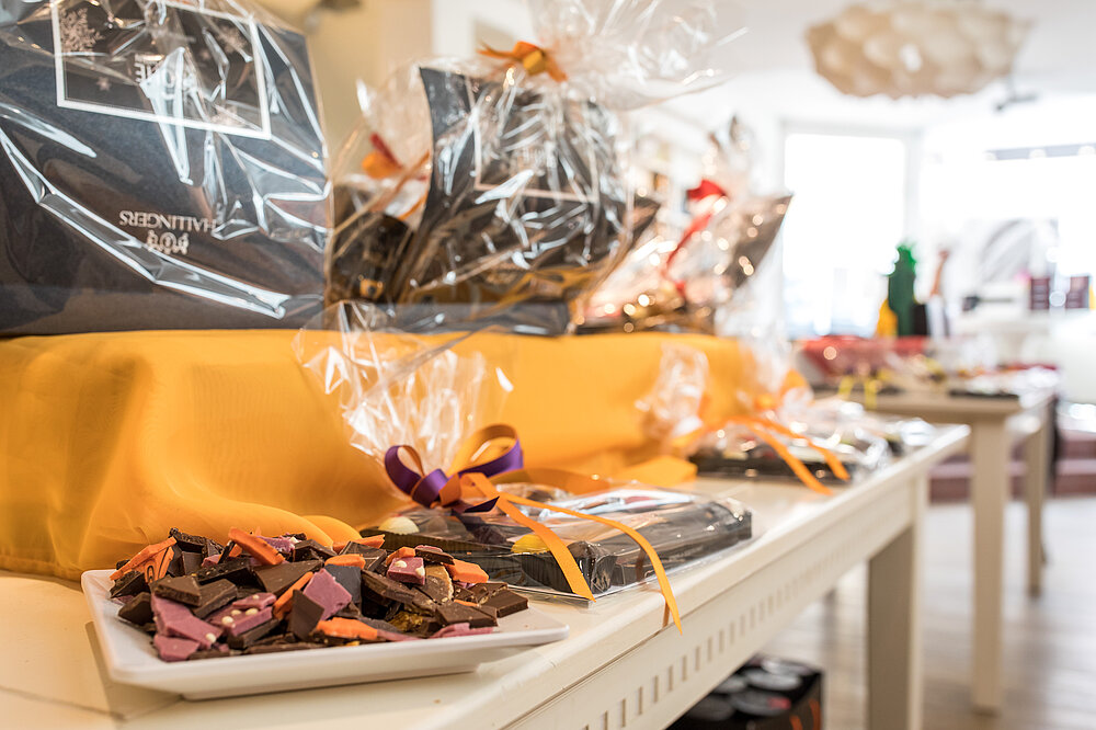 Das Bild präsentiert eine Verkostungstheke der Hallingers Genuss Manufaktur in Landsberg am Lech mit handgeschöpfter Schokolade, Serenity-Tafeln und Geschenken auf gelbem Tuch.