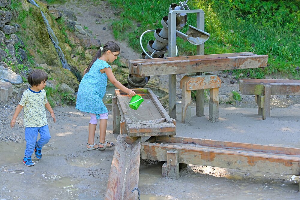 Ein Mädchen in einem blauen Kleid gießt Wasser aus einem grünen Eimer in eine hölzerne Wasserrinne, während ein kleinerer Junge daneben im nassen Sandbereich zuschaut. Im Hintergrund ist eine Archimedische Schraube aus Metall zu sehen.
