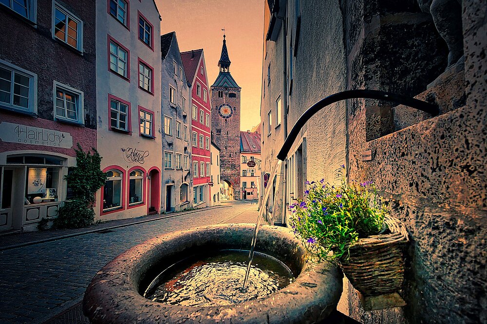 Albrecht Fietz' Foto zeigt den Trinkwasserbrunnen an der Alten Bergstraße in Landsberg am Lech mit sprudelndem Wasser, Lavendel und dem Schmalzturm im goldenen Sonnenuntergangshintergrund.