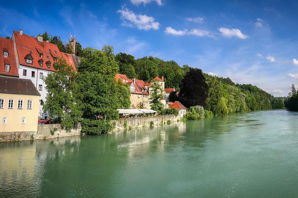Ansicht über den Fluss Lech auf eine historische Häuserzeile mit roten Dächern, einen Biergarten direkt am Wasser und den Jungfernsprungturm im Hintergrund unter blauem Himmel.