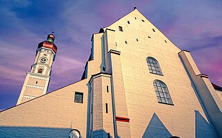 Abendlichtaufnahme der weißen Stadtpfarrkirche Landsberg am Lech mit rotem Turmhelm und gotischen Elementen vor purpurfarbenem Himmel.