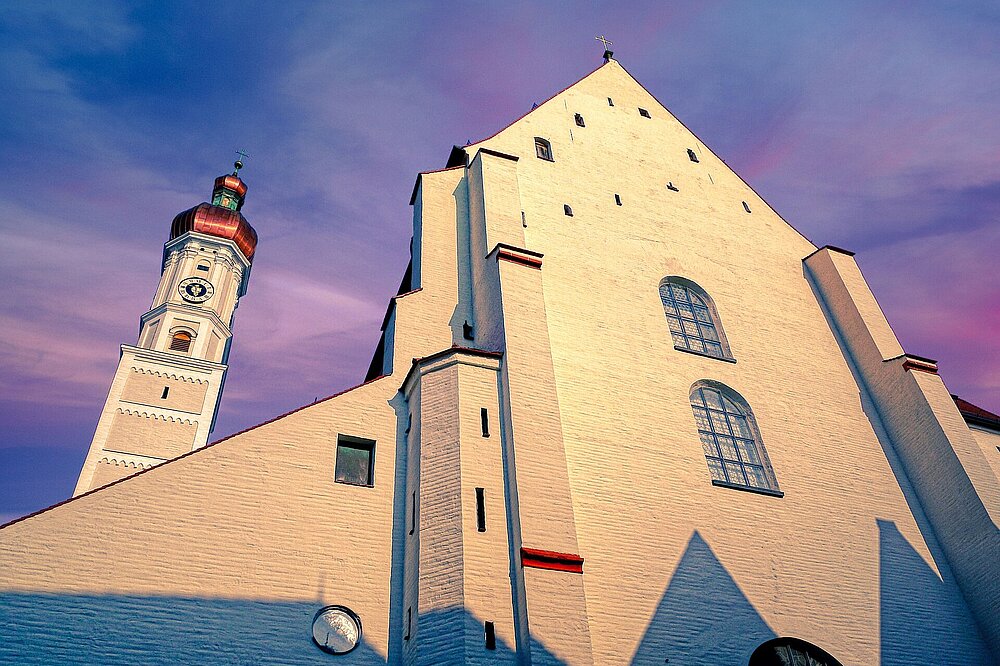 Sonnenuntergangsaufnahme der Stadtpfarrkirche Mariä Himmelfahrt Landsberg mit weißer Gotikfassade und rotem Turmhelm vor violettem Himmel.