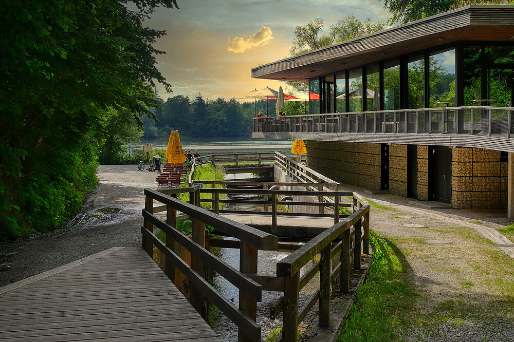 Die Teufelsküche ist ein modernes Gebäude mit großer Glasfront und Terrasse, das Restaurant Teufelsküche. Davor führt ein Holzsteg über einen kleinen Wasserlauf, im Hintergrund ist ein See (Lechstausee) unter einem gelblichen Sonnenuntergangshimmel zu sehen.