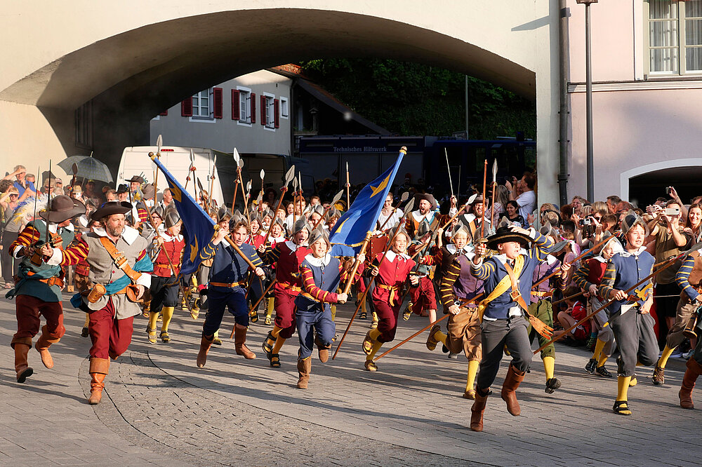 Ruethenfest-Umzug in Landsberg: Gruppe in Mittelalter-Kostümen (Rot, Blau, Gold) mit Speeren und Fahnen marschiert unter Bogen durch die Altstadtstraße.