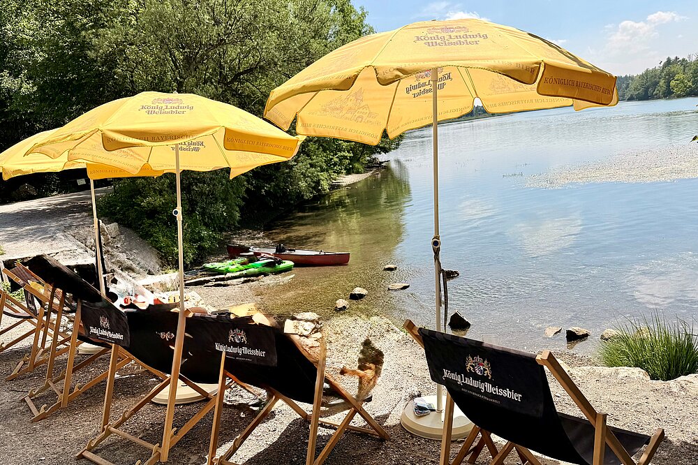 Mehrere Liegestühle unter gelben Sonnenschirmen der Marke König Ludwig Weissbier stehen auf einem Kiesstrand direkt am Ufer des Lechs. Im Hintergrund liegt ein rotes Kanu im flachen Wasser vor einem bewaldeten Ufer.