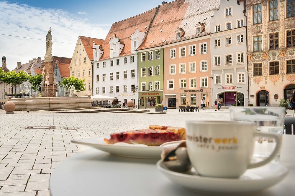 Café-Tisch mit Kaffeetasse, Wasserglas und einem Stück Kuchen im Vordergrund, im Hintergrund der historische Hauptplatz von Landsberg am Lech mit Brunnen, Bäumen, Pflastersteinen und farbigen, teils reich verzierten Fassaden mehrstöckiger Altstadthäuser bei leicht bewölktem Himmel.