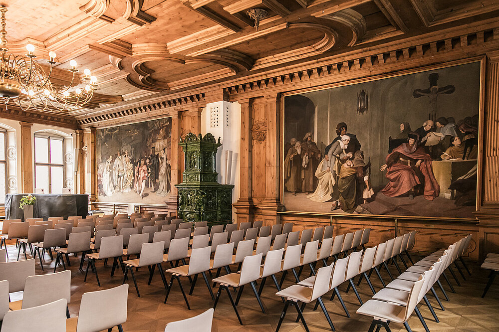 Festsaal im historischen Rathaus Landsberg am Lech: Historische Wandgemälde mit Szenen in barocker Holzpaneelung, grüner Kachelöfen, Kristalllüster und Stuhlreihen auf Parkettboden bei hellem Tageslicht.