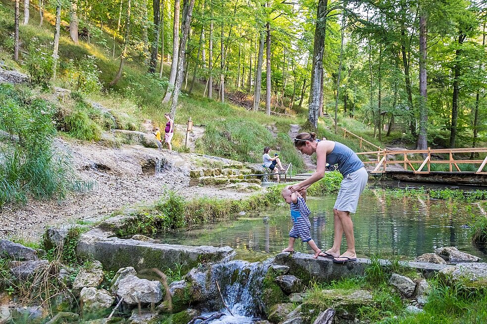 Eine Frau hält ein Kleinkind an den Händen, während es barfuß über Steine an einem kleinen Wasserlauf balanciert. Im Hintergrund sind weitere Personen an dem naturnahen Wasserspielbereich im Wald zu sehen.