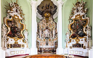 Rokoko‑Innenraum der Johanniskirche Landsberg: Prunkvoller Hochaltar mit Stuck, Fresken und Seitenaltären in hellgrünem Raum.