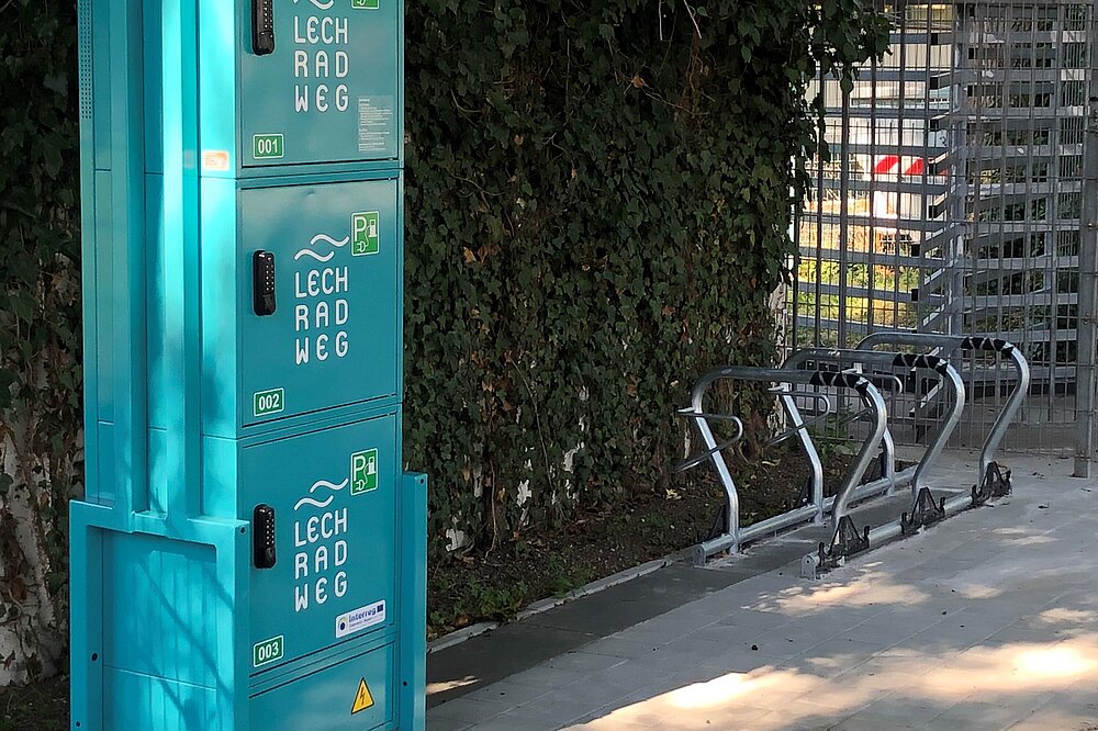 Blauer Ladepark für E-Bikes mit drei Stationen 'Lechweg P' neben Fahrradständern und Grünpflanzen – E-Bike Ladestation Lech-Radweg Landsberg am Lech Bayern (CC BY-NC Grün)