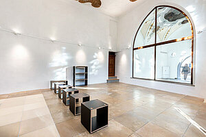 Helles Foyer im Klostereck Landsberg: Schwarze Möbelstapel (Hocker, Regale), hohes Rundbogenfenster mit Sonnenlicht, weiße Wände und Holzdeckendetails – minimalistischer Ausstellungsraum.