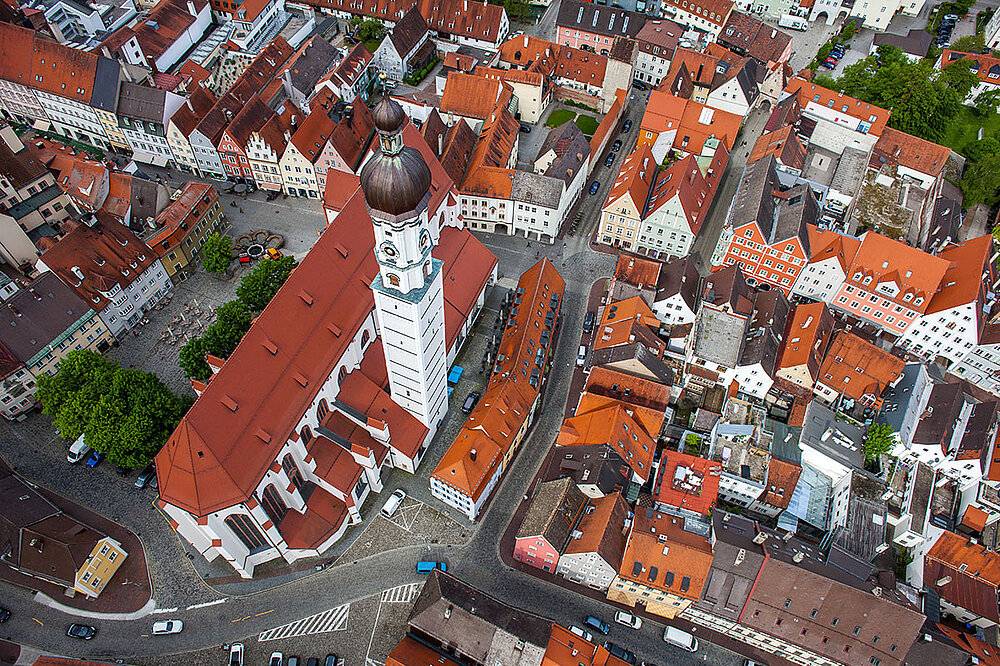Drohnenaufnahme der Stadtpfarrkirche Mariä Himmelfahrt Landsberg inmitten roter Dächern der Altstadt mit weißem Turm und Marktplatz.