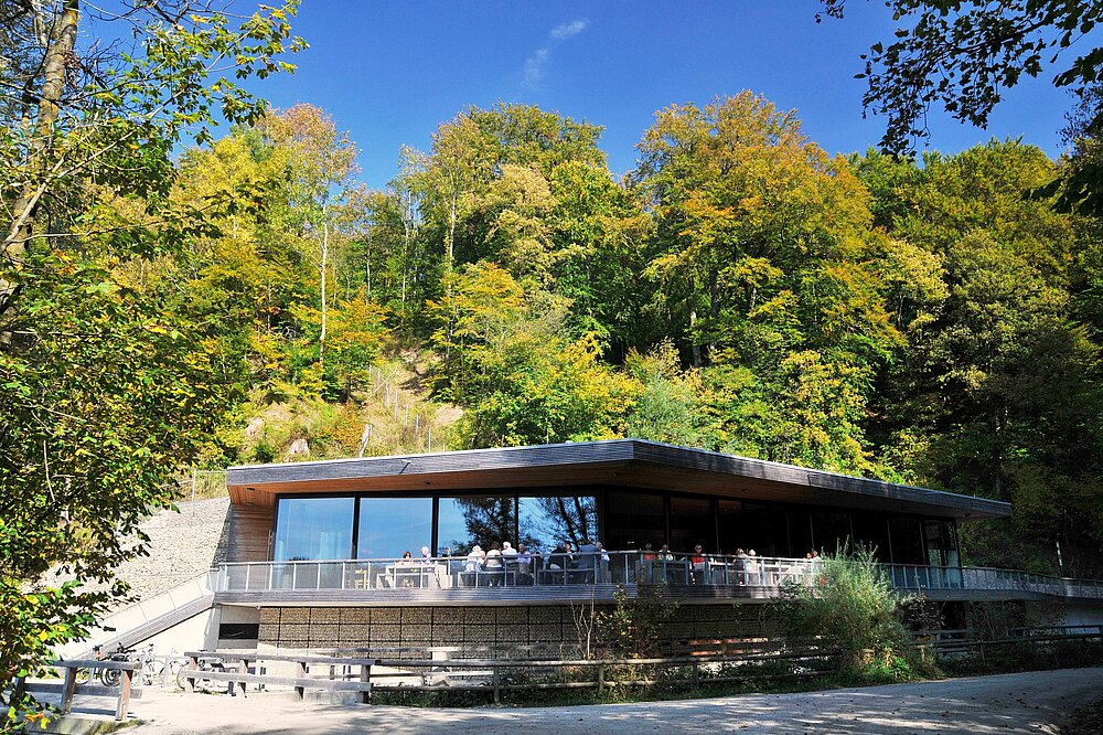 Das flache, moderne Gebäude des Restaurants Teufelsküche mit einer breiten, verspiegelten Glasfront und einer Außenterrasse, auf der Gäste sitzen. Das Gebäude liegt direkt am Fuß eines steilen Hanges, der dicht mit herbstlich gefärbten Bäumen bewachsen ist, unter blauem Himmel.