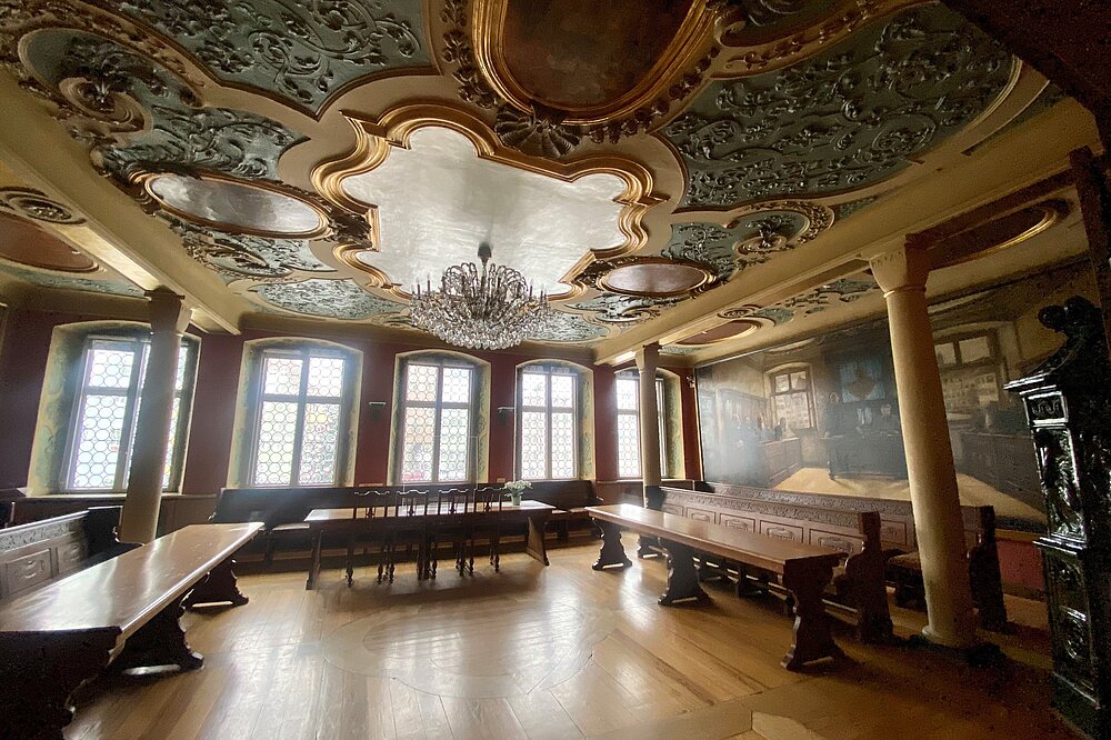 Herkomer-Saal im historischen Rathaus Landsberg am Lech: Prächtige barocke Deckenstukkaturen mit Stuckreliefs und Lüster über rustikalen Eichentischen, bemalten Wänden und bleiverglasten Fenstern bei warmem Sonnenlicht.