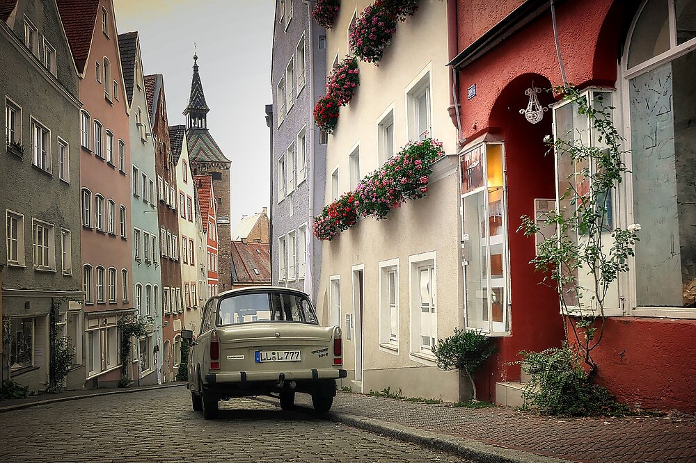 Albrecht Fietz' Foto fängt die steile Alte Bergstraße in Landsberg am Lech ein, mit dem Schmalzturm im Hintergrund, bunten Fachwerkhäusern, Blumenkästen und einem Oldtimer-Auto auf Kopfsteinpflaster.