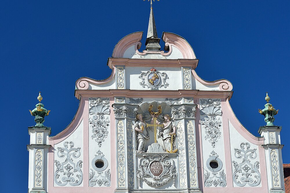 Barocker Giebel des historischen Rathauses Landsberg am Lech: Stuckreliefs mit allegorischen Figuren, Wappen, Girlanden und Wetterfahne auf pastellrosa Fassade vor blauem Himmel.