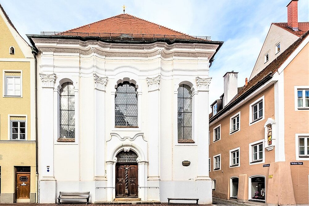 Weiße Rokoko‑Fassade der Johanniskirche Landsberg neben pastellfarbenen Altstadthäusern mit Bänken und Eingang.
