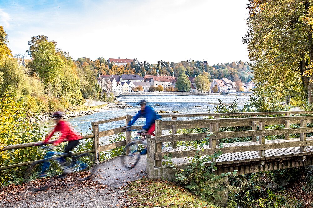 Radfahrer in Rot und Blau fahren über Holzbrücke mit Herbstlaub gegenüber Lechwehr und Altstadt Landsberg am Lech – Radtouren Lech Bayern im Herbst (CC BY-SA Klinger)