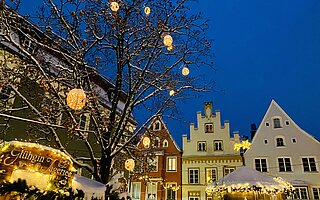 Christkindlmarkt auf dem Hauptplatz Landsberg: Weihnachtsbaum, beleuchtete Stände mit Glühwein und Deko vor dem Rathaus und Fachwerkhäusern bei Schneefall.